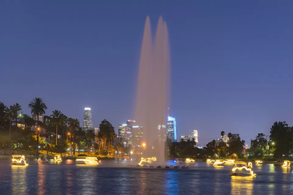 View of Echo Park Lake at night with artesian fountain and illuminated swan paddle boats and Los Angeles skyline in background